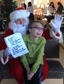 santa waves and holds a christmas list with a student on his lap